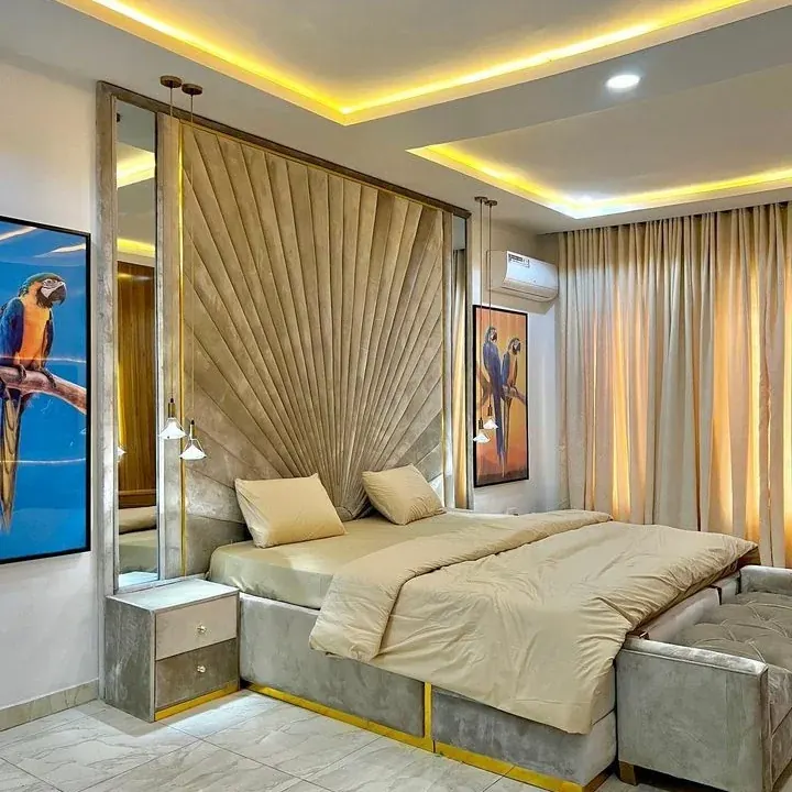 A finished apartment bedroom with a wide upholstered bed and a gold fan-panel headboard wall under a lit ceiling