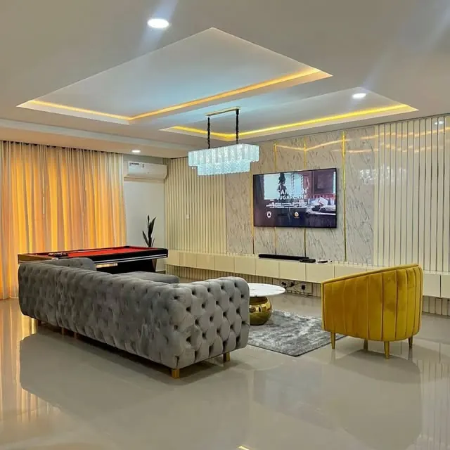 A finished living room with a grey button-tufted sofa, a yellow armchair, a lit tray ceiling and a TV wall