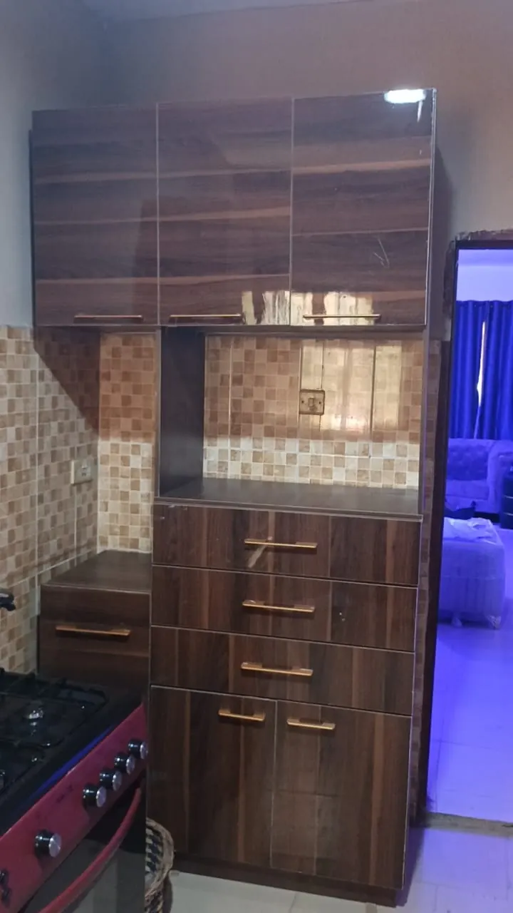 A tall wood-grain kitchen unit with glossy upper cabinets, a recess for a microwave, drawers with gold handles and a cooker beside it