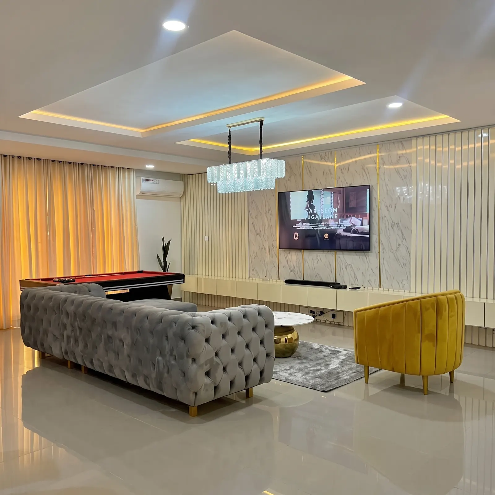 A grey button-tufted chesterfield sofa and a mustard chair in a finished living room with a pool table and a lit TV wall