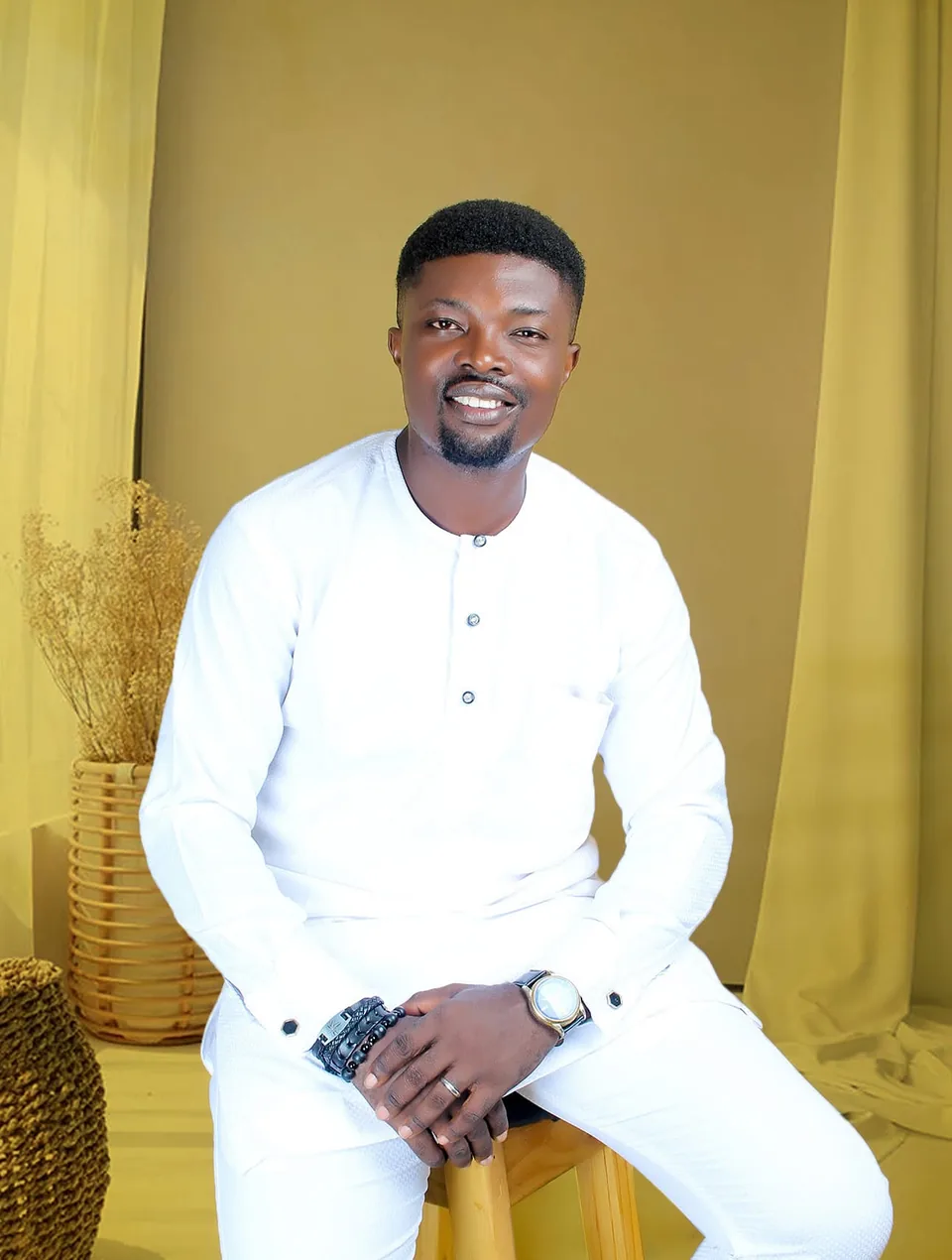 Kolawole, the owner, smiling, seated in a white outfit against a mustard backdrop