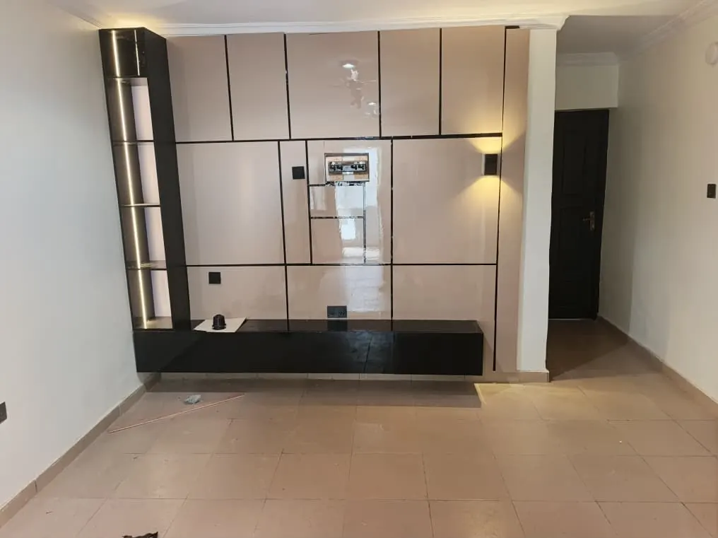 A finished living-room TV wall with cream gloss panels in black frames, a lit shelf column on the left and a long floating black console