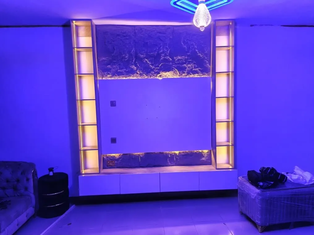 A TV wall at night with glowing LED shelf columns either side of a dark mirrored panel, under blue ceiling light