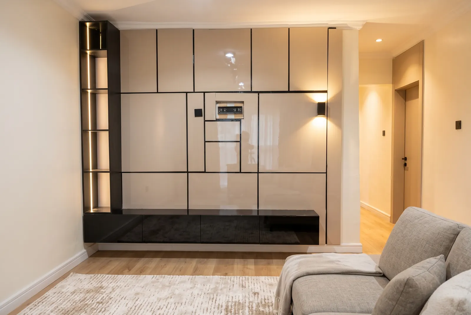 A TV wall of glossy cream panels with thin black trim, a lit shelf column on the left, a recessed TV niche and a floating black console, in a warm living room